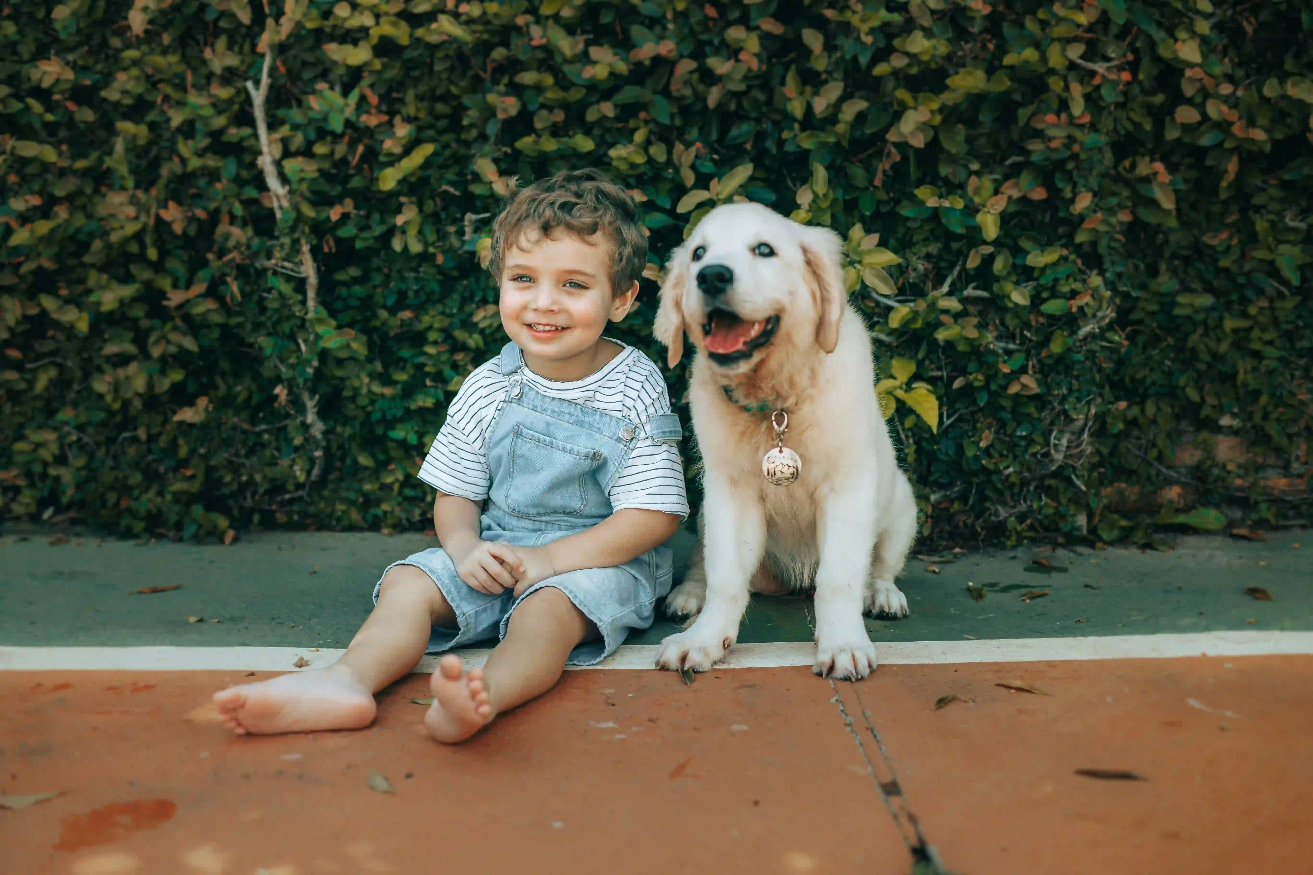 A young child sitting side by side with a golden retriever puppy outdoors, both smiling toward the camera