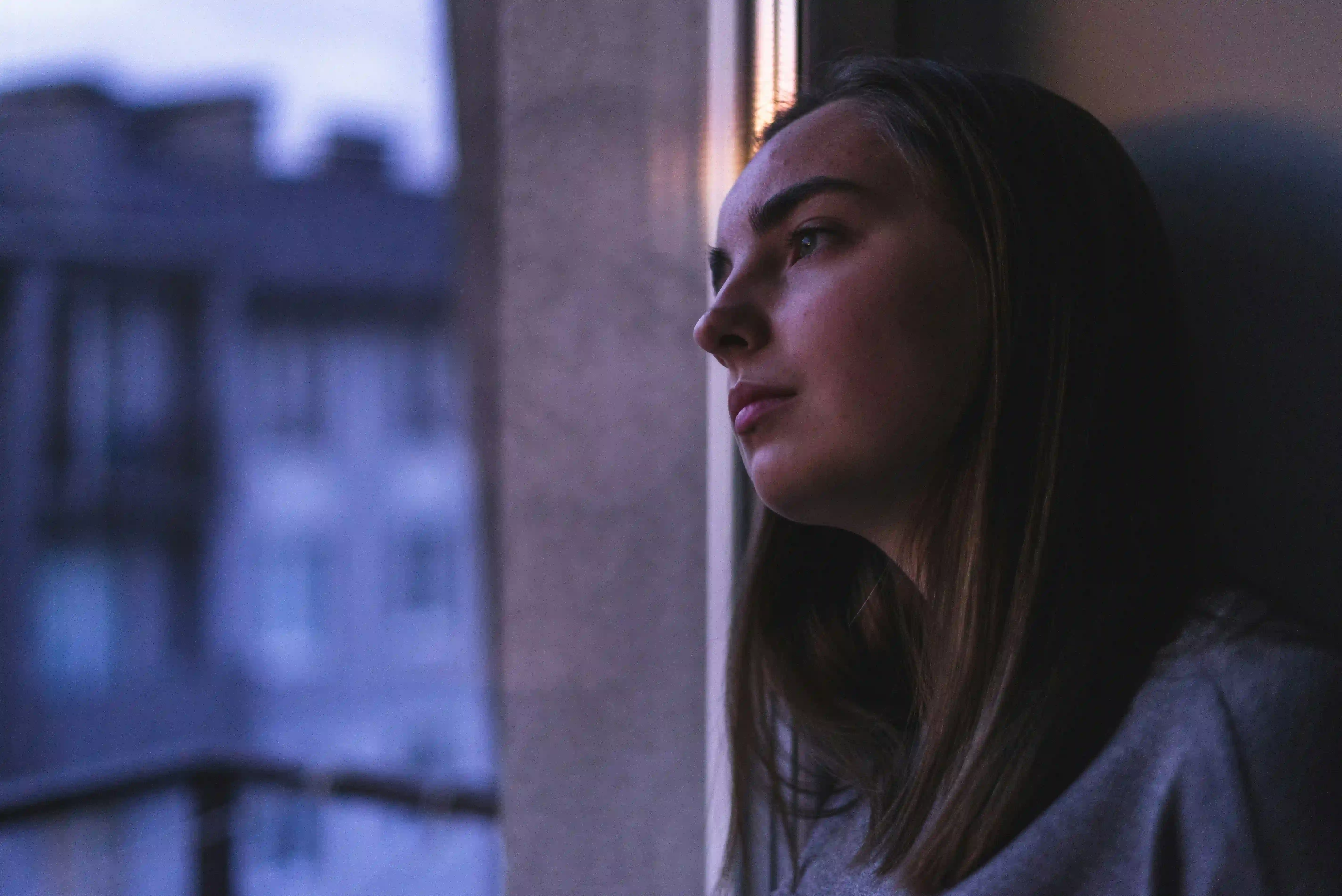 A woman gazing quietly out a window at dusk, city rooftops visible in the soft blue evening light