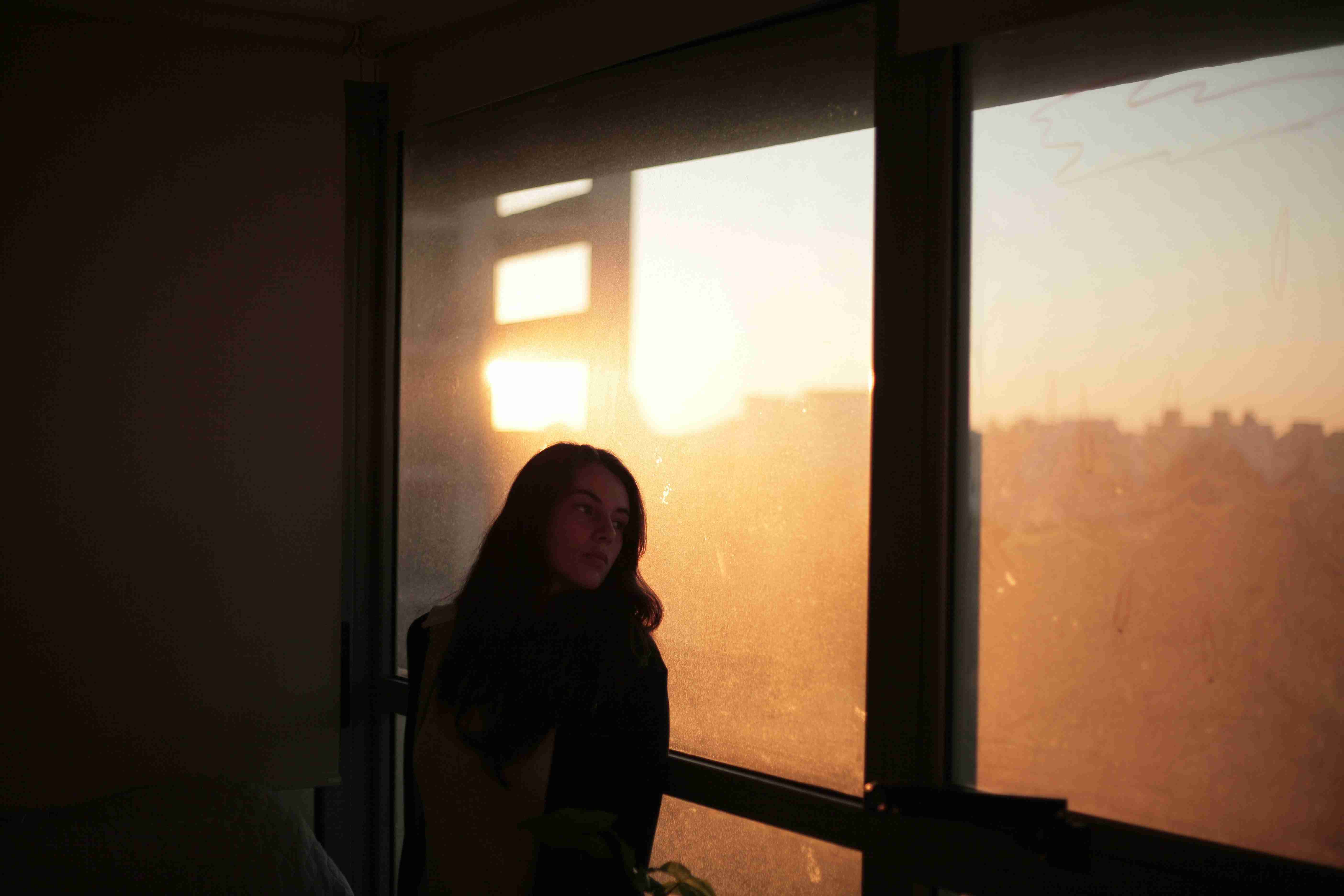 A woman standing quietly by a window at dusk, looking out in a moment of stillness