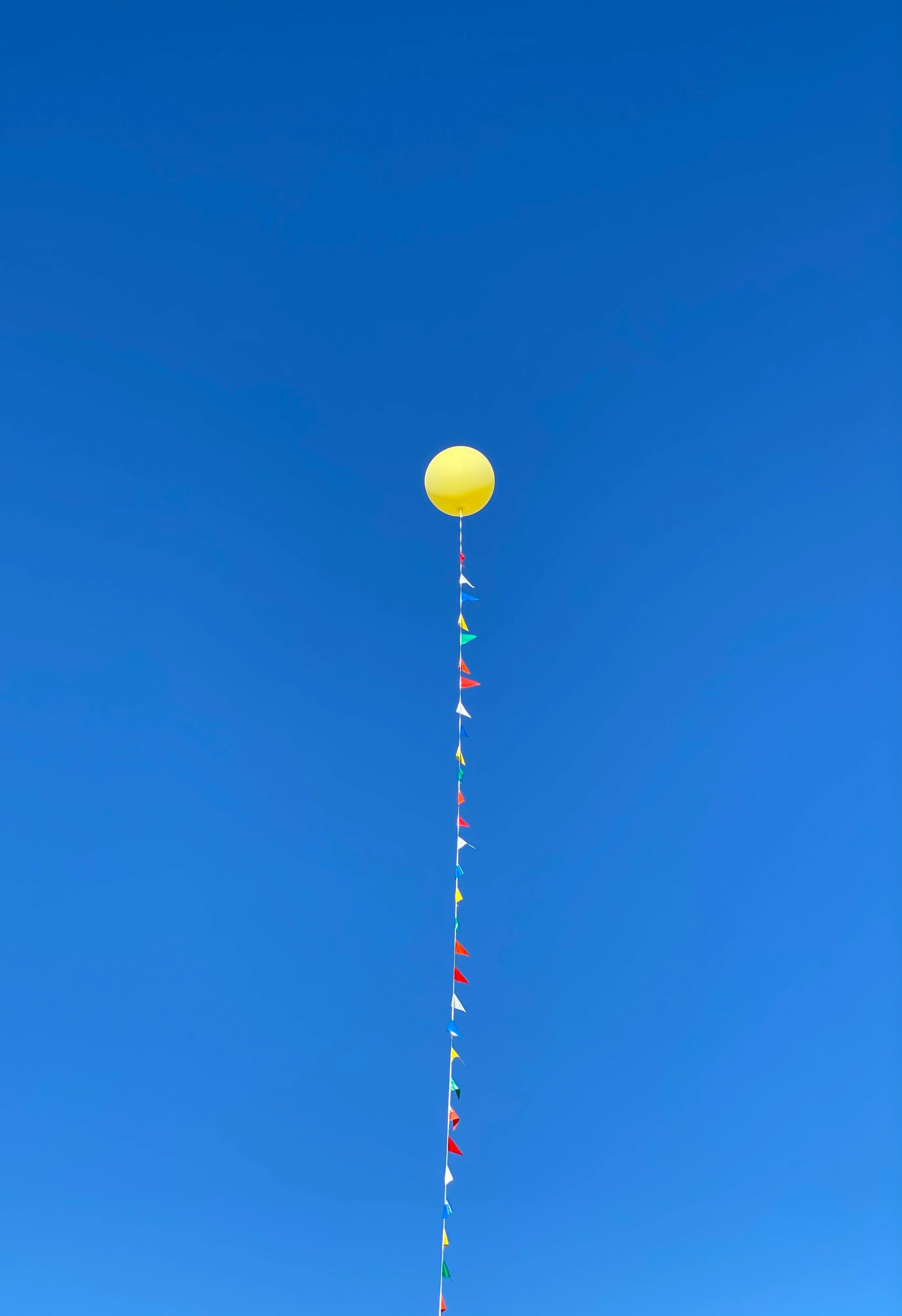 A single yellow balloon with a string of small colourful flags rising against a clear blue sky